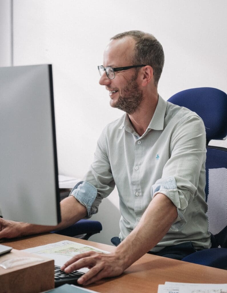 A portrait of a smiling man with a short beard and glasses sitting at a desk and working on a computer monitor. He is wearing a light greenish-grey patterned button-down shirt with rolled-up sleeves, and architectural plans are lying on the desk in front of him.