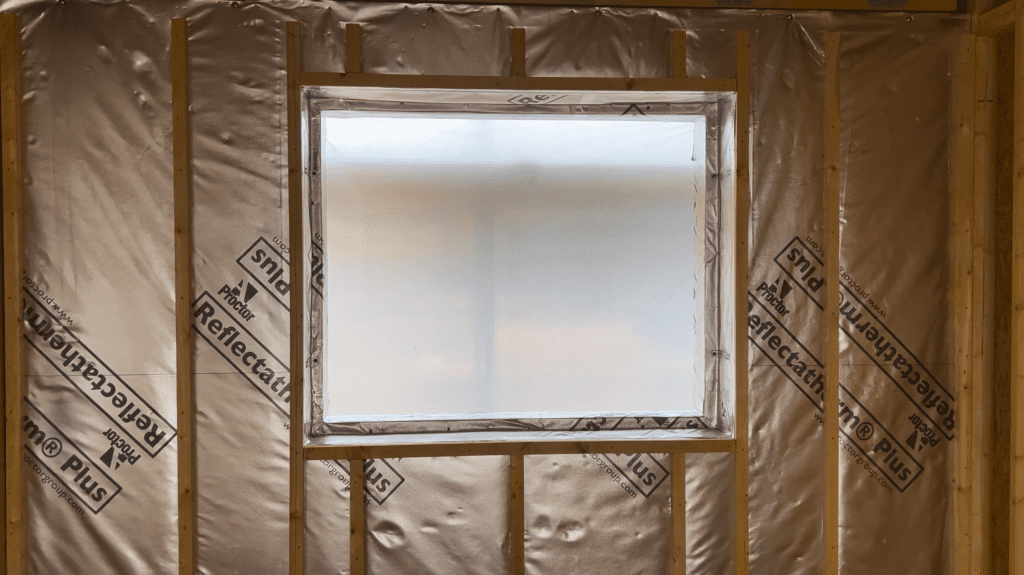 An interior view of a timber frame wall under construction, framing a rectangular window opening. The entire inner face of the wall is covered with a highly reflective silver foil membrane, printed with the repeating logo "Proctor Reflectatherm Plus".
