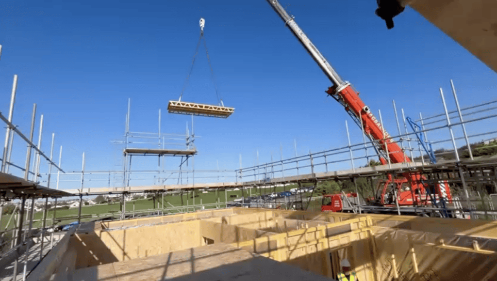 A construction site under a clear blue sky where a large red crane is lowering a prefabricated timber floor or roof panel onto the wooden framework of a building surrounded by metal scaffolding.