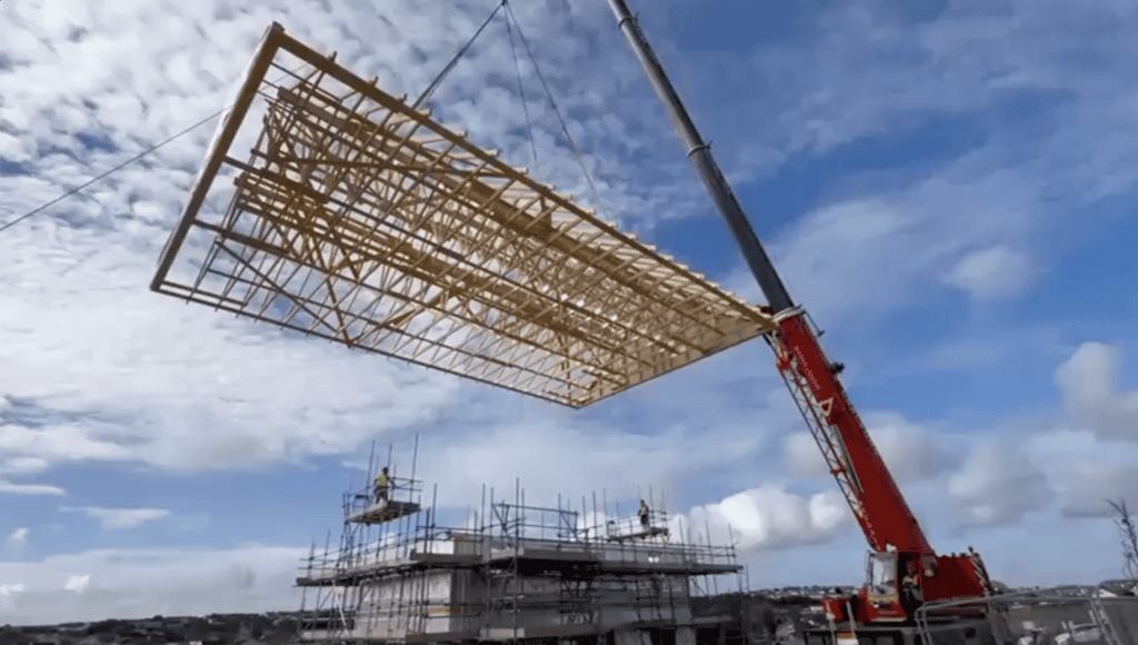 A massive prefabricated wooden roof truss framework being hoisted high into the air by a red crane on a construction site. Scaffolding and construction workers are visible below against a cloudy blue sky.