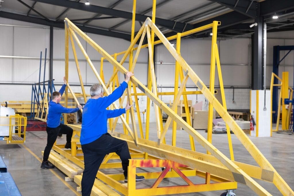 Two workers in blue long-sleeve shirts inside a large factory assembling a large, yellow-painted wooden roof truss. One worker is stepping up onto the base of the frame to position a piece of timber.