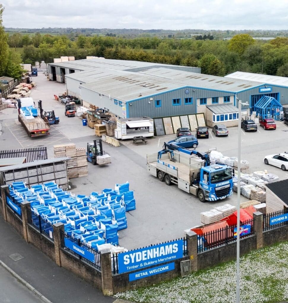 An elevated view of a sprawling Sydenhams Timber & Builders Merchants yard. The site features a long grey warehouse with blue trim, extensive stacks of timber and paving slabs, a large cluster of blue bulk bags, a small food truck, forklifts, and a blue Sydenhams flatbed lorry equipped with a crane.