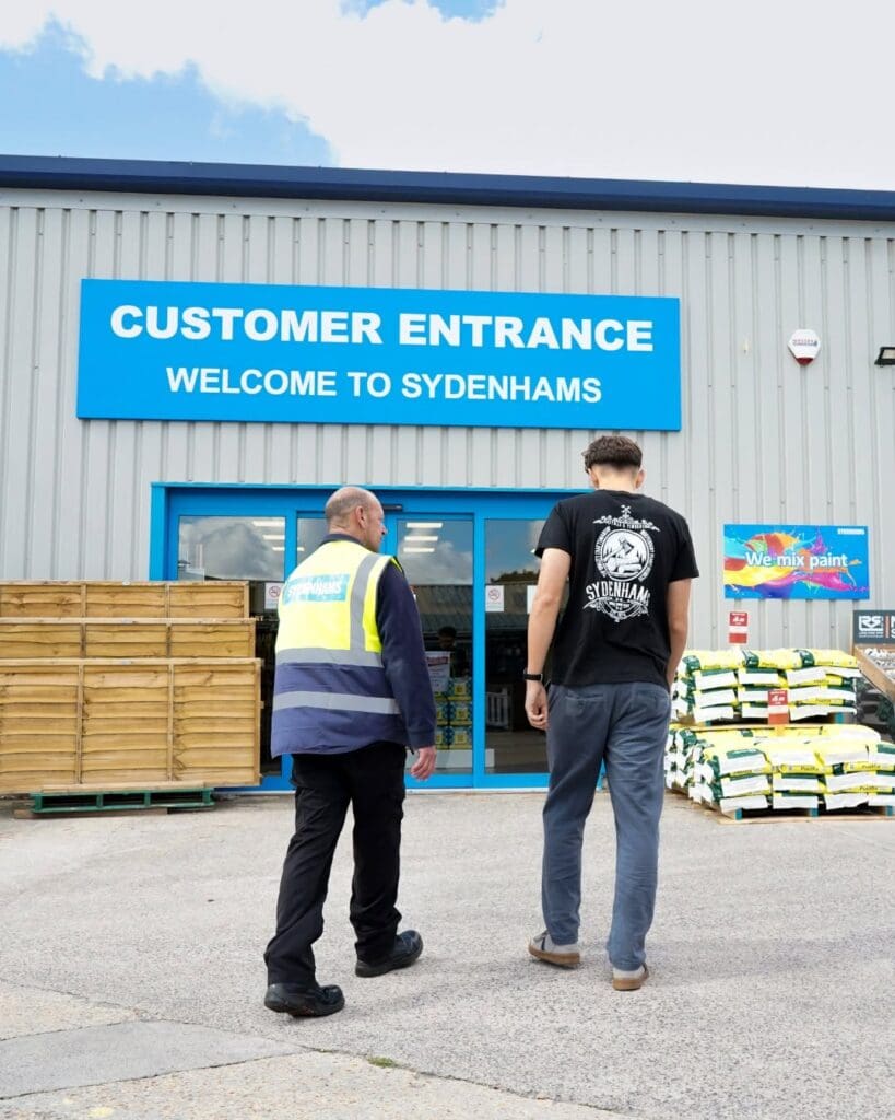 A rear view of two men walking toward the automatic glass doors of a building. A large blue sign above the entrance reads "CUSTOMER ENTRANCE WELCOME TO SYDENHAMS". One man wears a high-visibility Sydenhams vest, and the other wears a black t-shirt featuring a white vintage-style Sydenhams logo on the back.