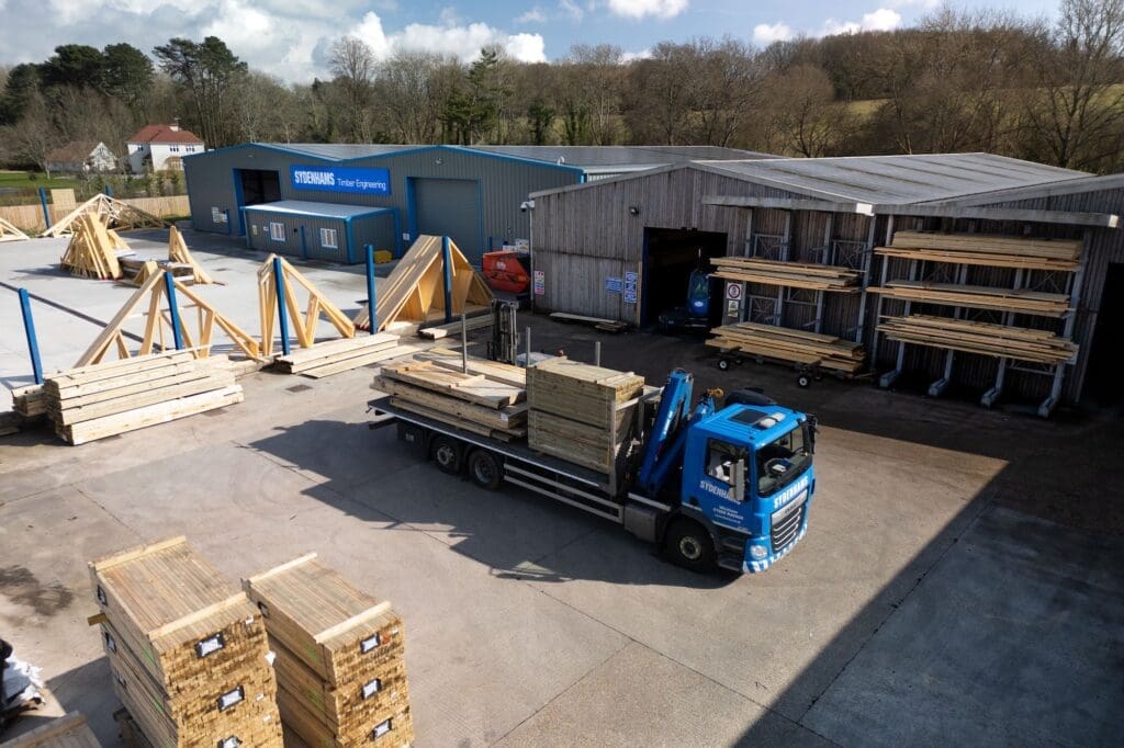 An elevated view of a Sydenhams Timber Engineering yard. A blue flatbed lorry loaded with stacks of timber is parked in the center. The yard contains multiple assembled wooden roof trusses, stacks of lumber in storage racks, and large industrial buildings in the background.