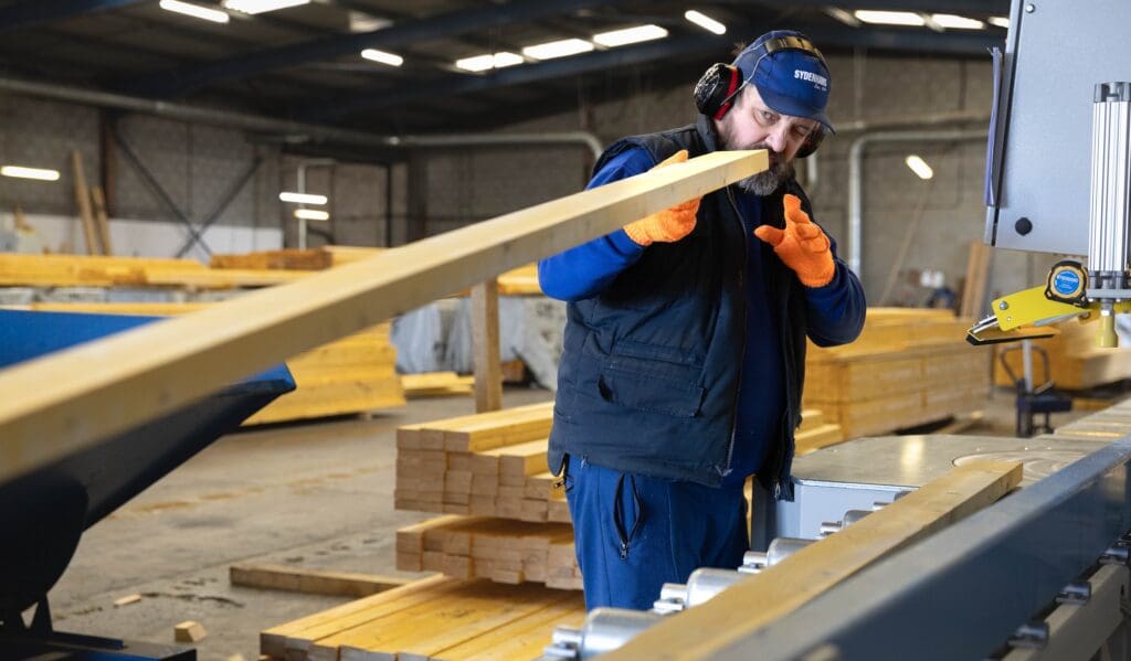 A factory worker wearing a blue Sydenhams cap, ear defenders, a dark vest, and bright orange grip gloves is holding a long piece of timber, carefully feeding it into an industrial cutting or milling machine inside a warehouse.