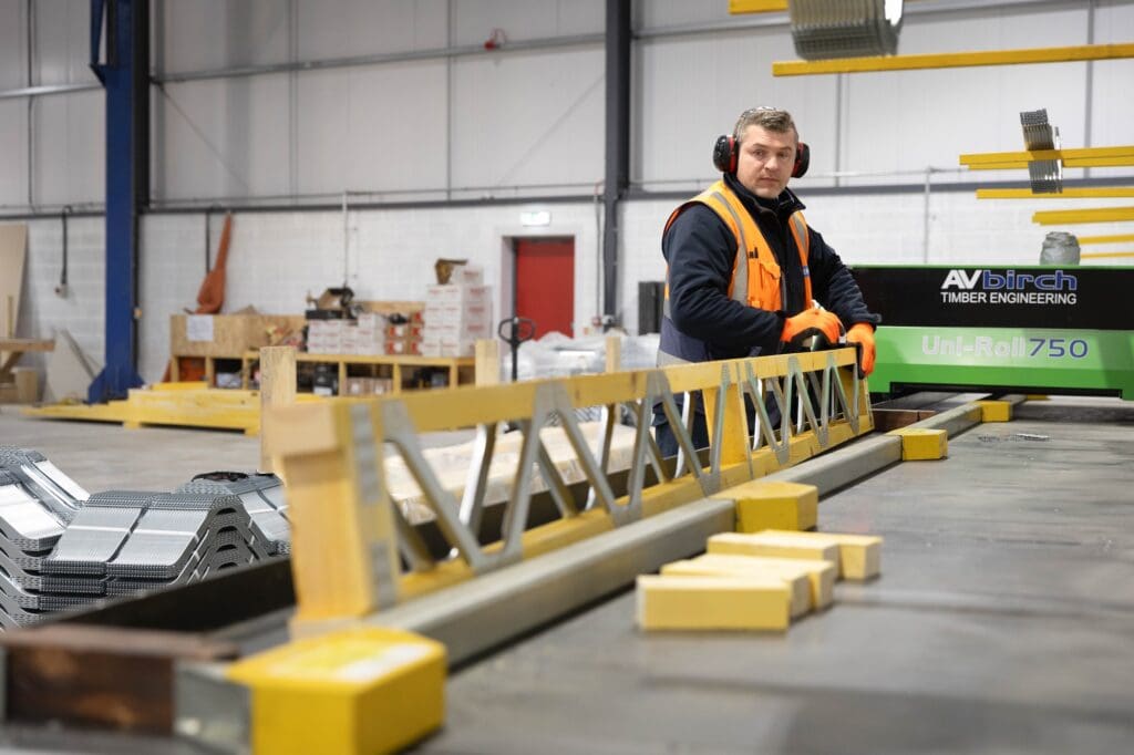 A factory worker wearing ear defenders, safety glasses, a high-visibility vest, and orange gloves stands at a large assembly table, handling a prefabricated timber metal web joist. A green "AV Birch Timber Engineering Uni-Roll 750" machine is visible in the background.