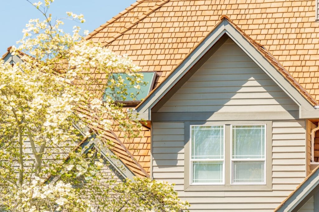 The exterior of a house featuring light grey horizontal siding, white-framed windows, and a roof made of wooden shingles with a flat skylight. A blooming tree with white flowers partially obscures the view in the foreground.